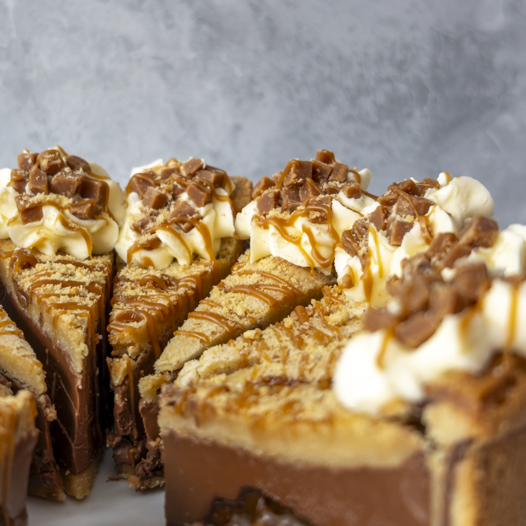 Close-up of several slices of a Banoffee Cookie Pie, topped with whipped cream, chocolate pieces, and caramel drizzle, set against a soft gray background.