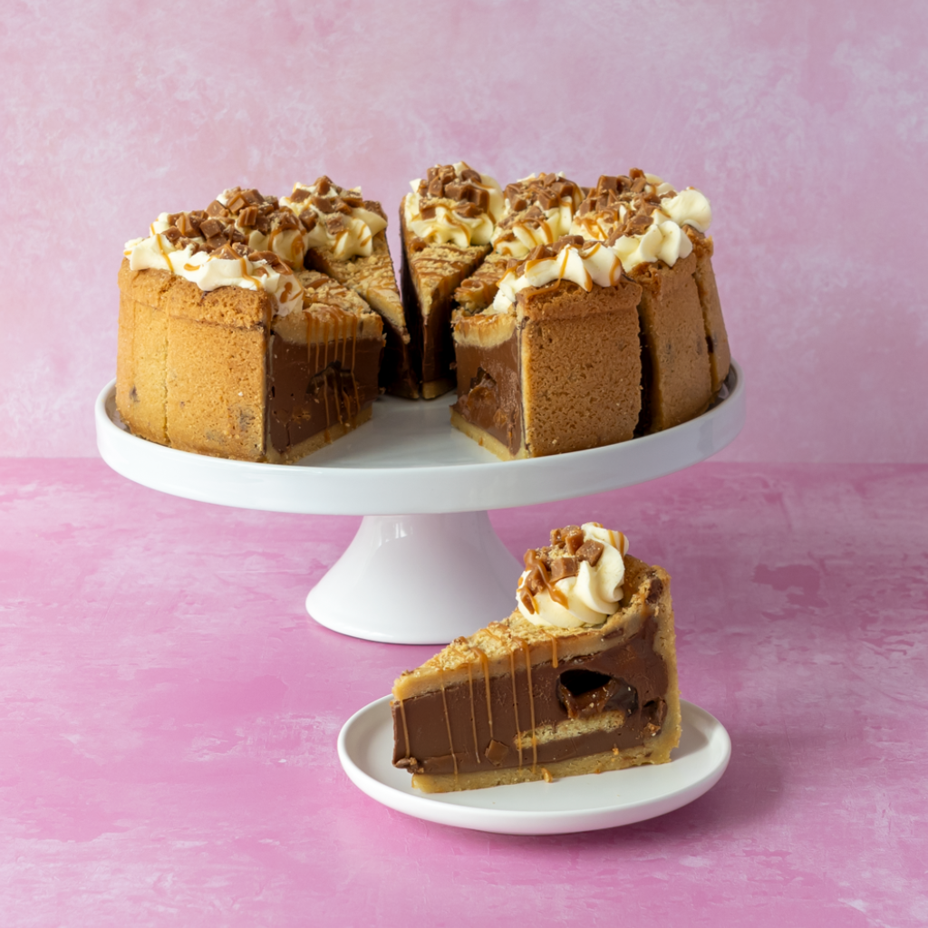A layered Full Banoffee Cookie Pie Slice with cream and chocolate drizzle, shown on a white cake stand with a single slice on a small plate in front, set against a pink textured background.
