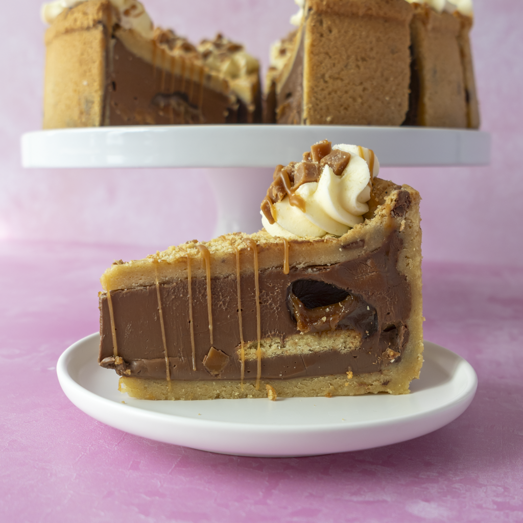 A slice of chocolate and caramel pie with a biscuit crust, whipped cream, and caramel drizzle, served on a white plate in front of a whole pie on a white cake stand against a pink background.