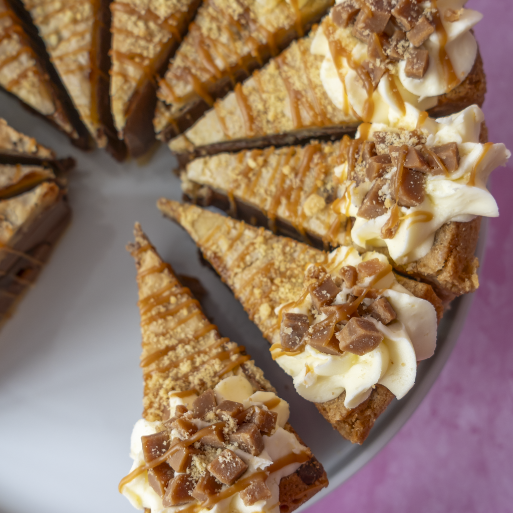 A close-up of a sliced Banoffee Cookie Pie Slice with whipped cream, chocolate drizzle, crushed candy bars, and a crumb topping, served on a plate against a purple background.