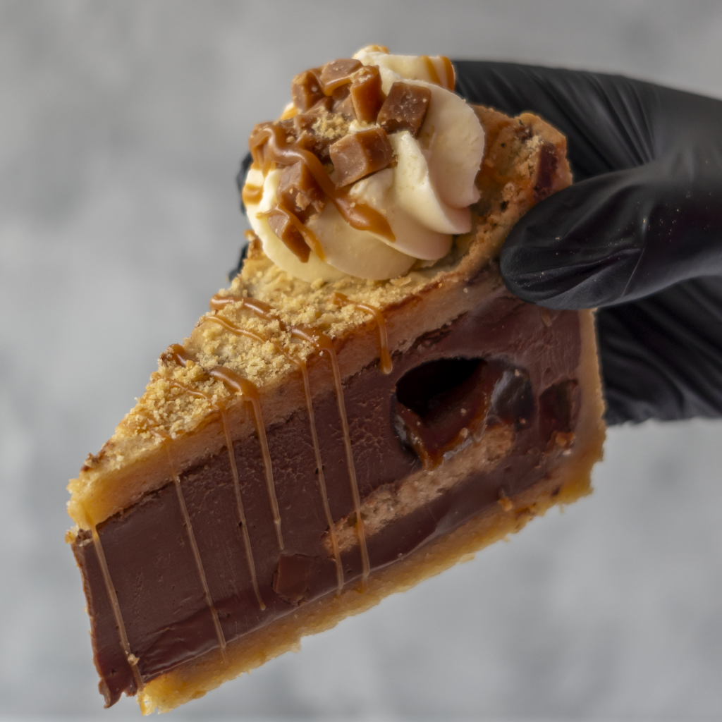 A close-up of a hand wearing a black glove holding a slice of chocolate cookie pie with caramel drizzle, whipped cream, and chocolate chunks on top, against a soft gray background.