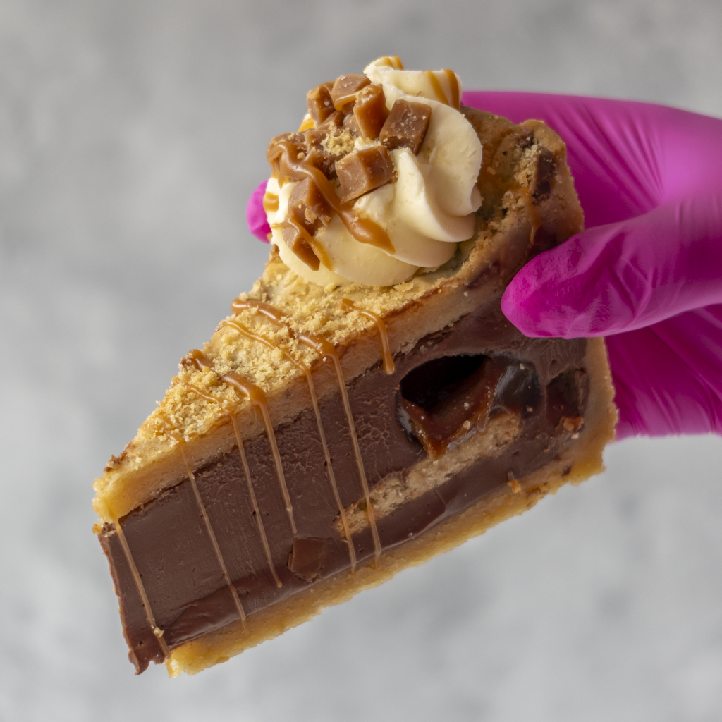 A close-up of a hand wearing a bright pink glove holding a slice of Banoffee Cookie Pie Slice with whipped cream, caramel drizzle, and candy pieces on top, against a soft gray background.