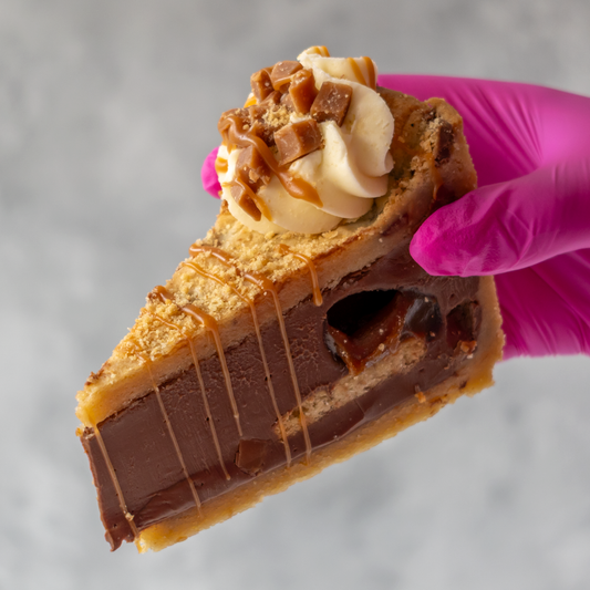 A close-up of a hand wearing a bright pink glove holding a slice of Banoffee Cookie Pie Slice with whipped cream, caramel drizzle, and candy pieces on top, against a soft gray background.