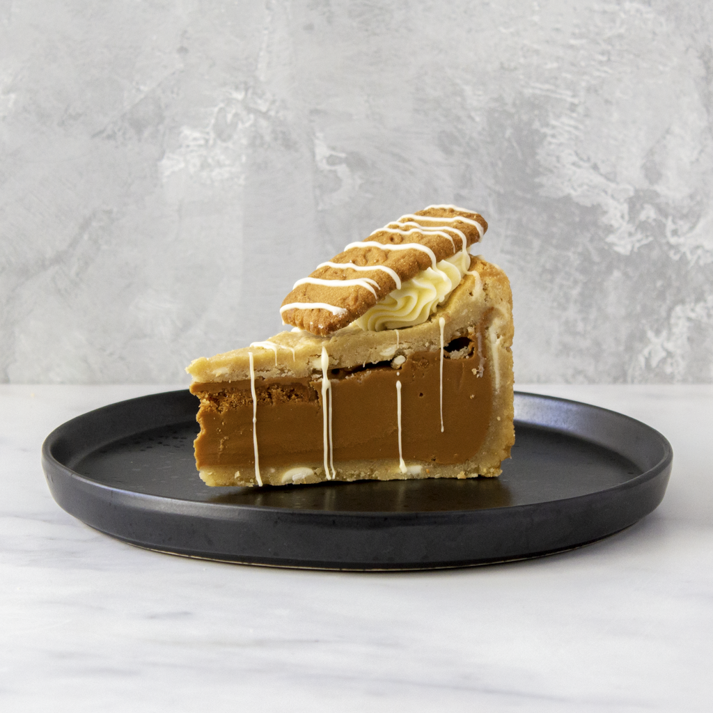 A slice of Cookie Pie with Biscoff on a black plate, featuring a golden-brown pastry crust, a thick caramel-like filling, piped cream filling, and a biscuit on top with white drizzle, set against a grey textured background.