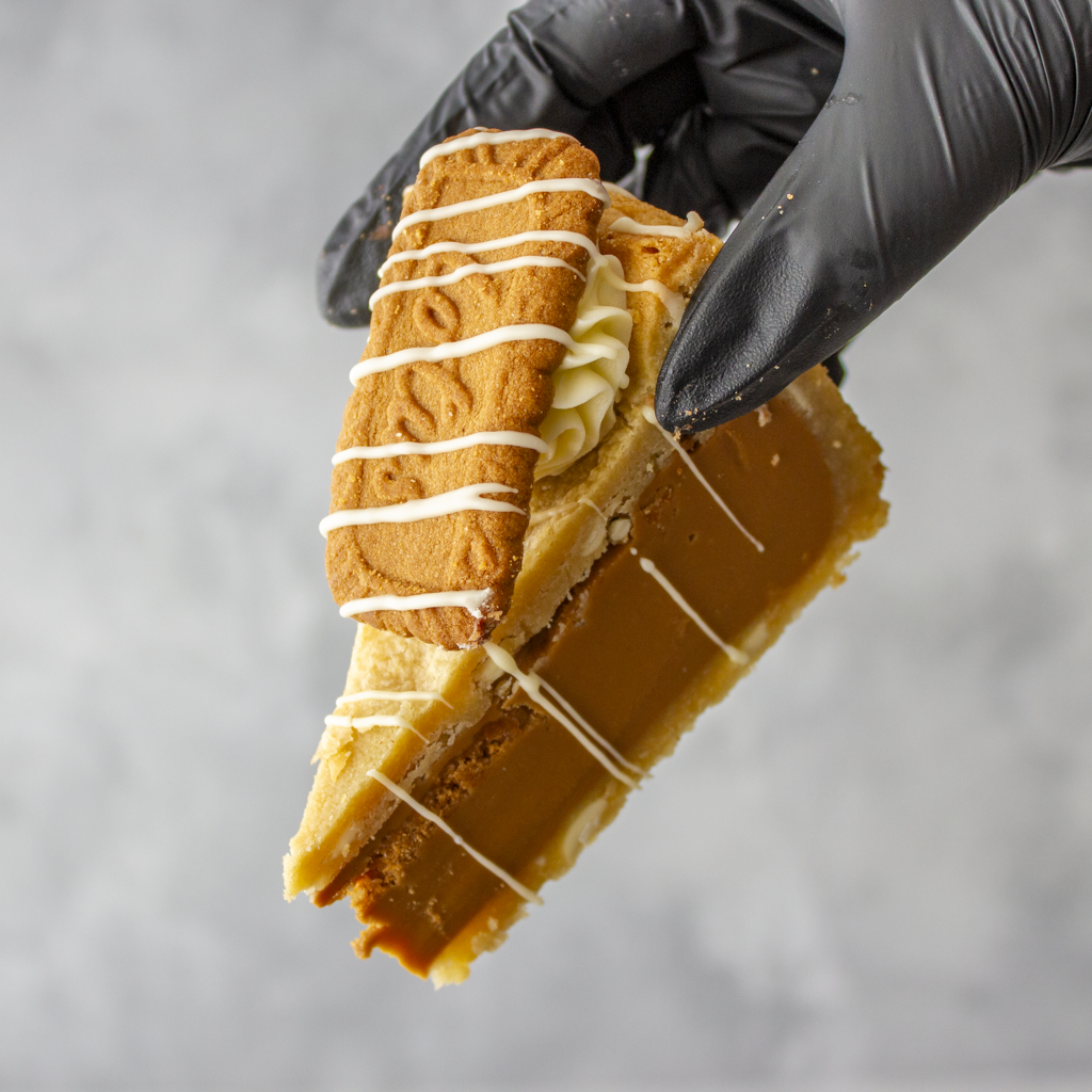 A hand wearing a black glove holds a slice of Cookie Pie with Biscoff featuring caramel-like custard, a biscuit, whipped cream, and white chocolate drizzle against a neutral grey background.