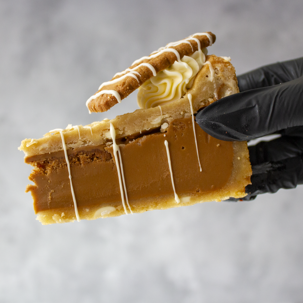 A close-up of a slice of Cookie Pie with Biscoff held by a hand wearing a black glove, topped with a cream swirl and a biscuit, against a soft grey background.