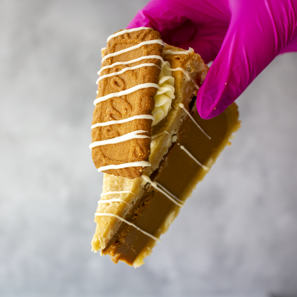 A hand wearing a bright pink glove holds a large slice of Cookie Pie with Biscoff, likely a caramel and cookie pie or cake, topped with white chocolate drizzle and a biscuit or cookie layer, against a soft grey background.