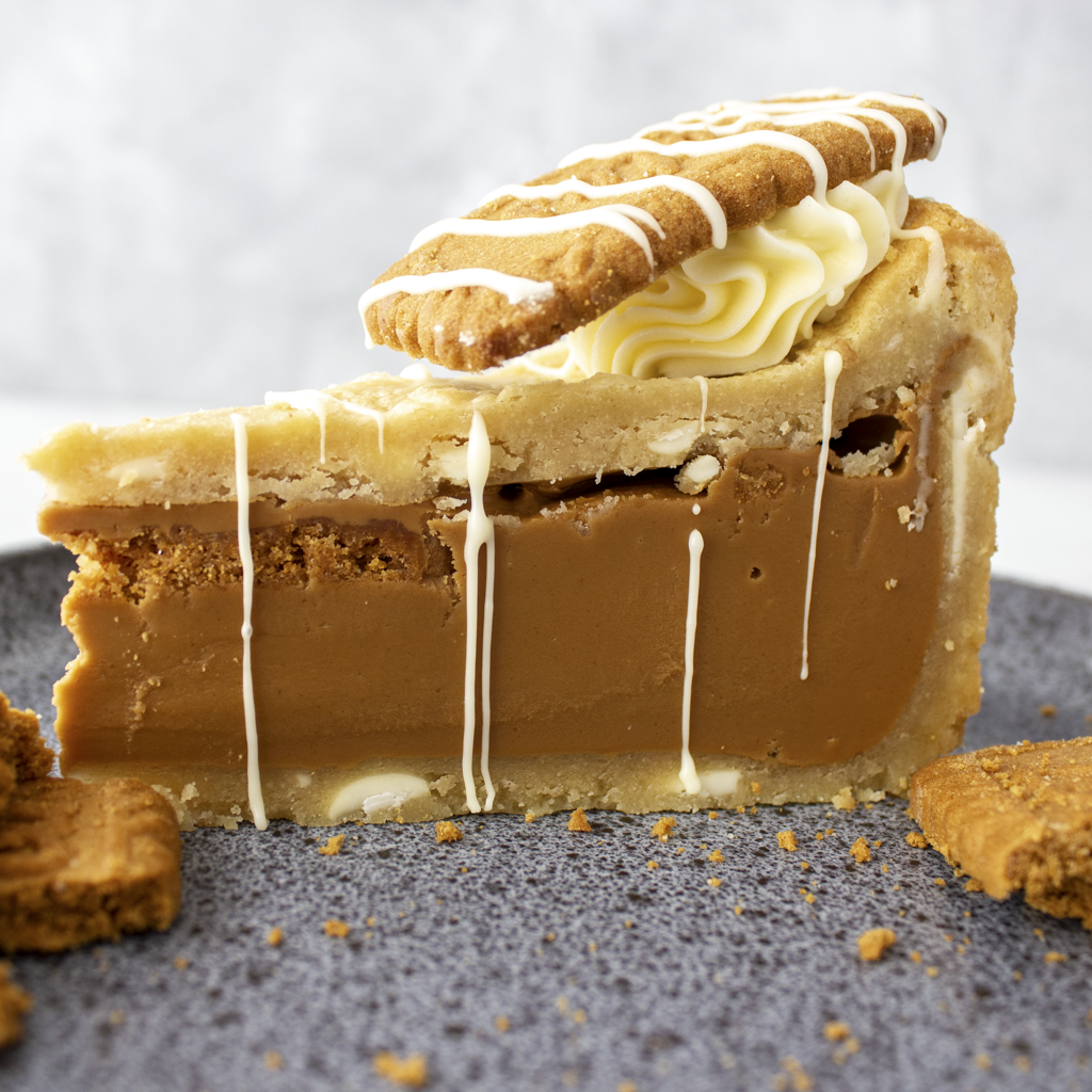 A close-up of a slice of Cookie Pie with Biscoff with a thick caramel-like filling, biscuit crust, piped cream swirl, and a cookie on top, set on a dark textured plate.