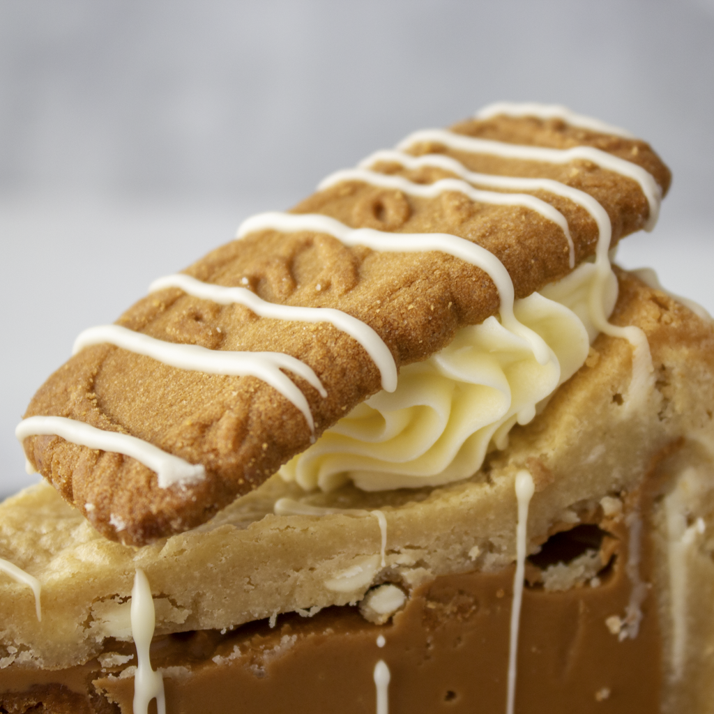 A close-up of a cookie pie slice with Biscoff with a golden-brown cookie on top, white drizzle, pale cream filling, and a caramel-brown layer underneath, photographed against a soft light grey background.