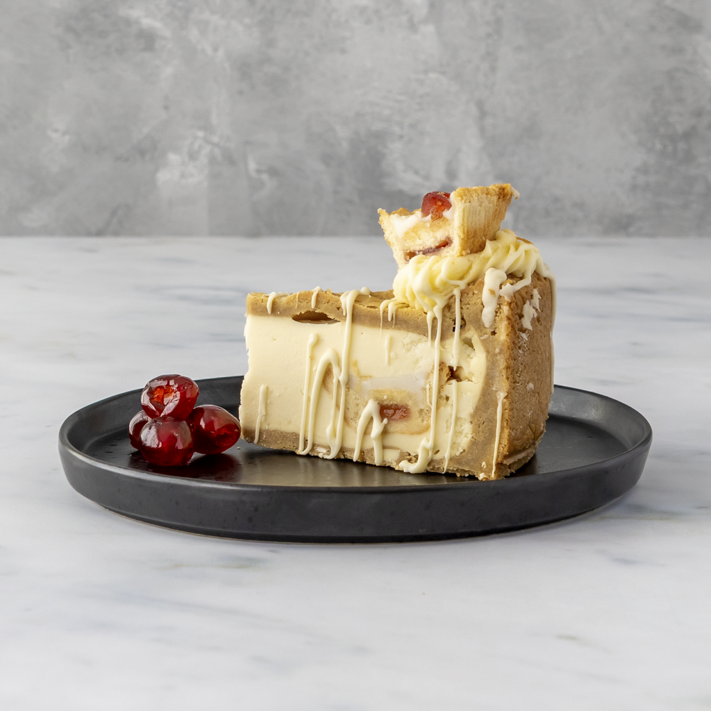 Slice of DoughGirl Cherry Bakewell Cookie Pie with white drizzle on a black plate, surrounded by red cherries, against a gray background.