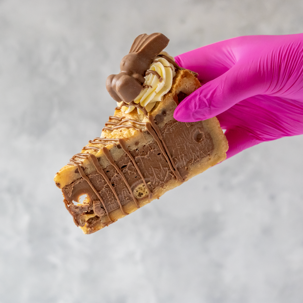 Hand holding a slice of Easter dessert with Cookie Pie and Malteser chocolate bunny, against a neutral background.