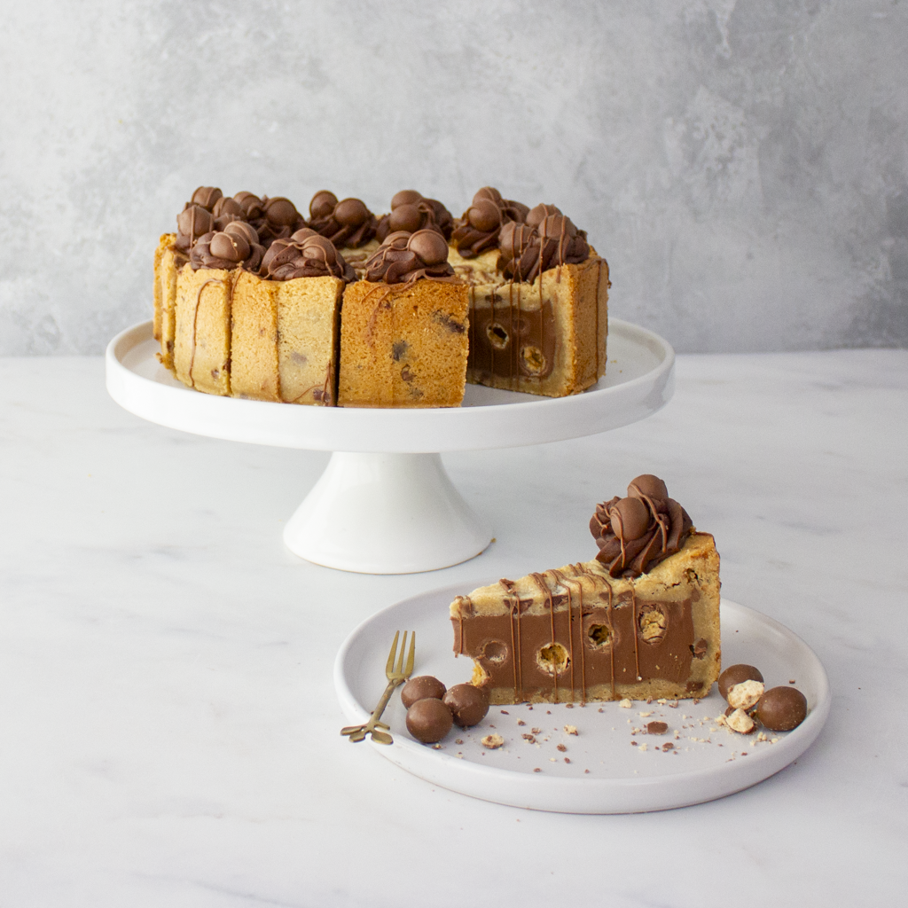 Chocolate chip cookie dough dessert with a slice on a plate, on a white cake stand against a gray background.