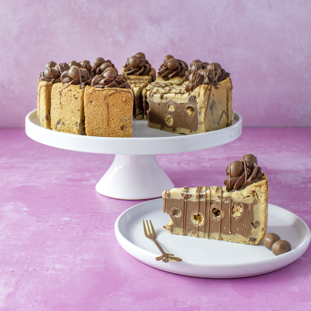 DoughGirl Full Malteser Cookie Pie on a white cake stand with a slice on a plate against a pink background.