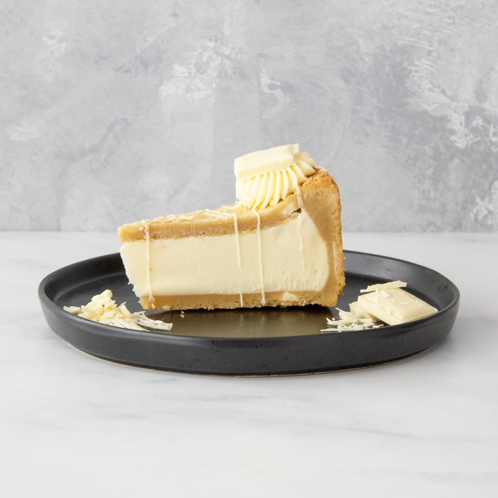 A slice of creamy cookie pie with Milkybar with a graham cracker crust, topped with white cream and drizzled with chocolate, served on a round black plate against a grey textured background.