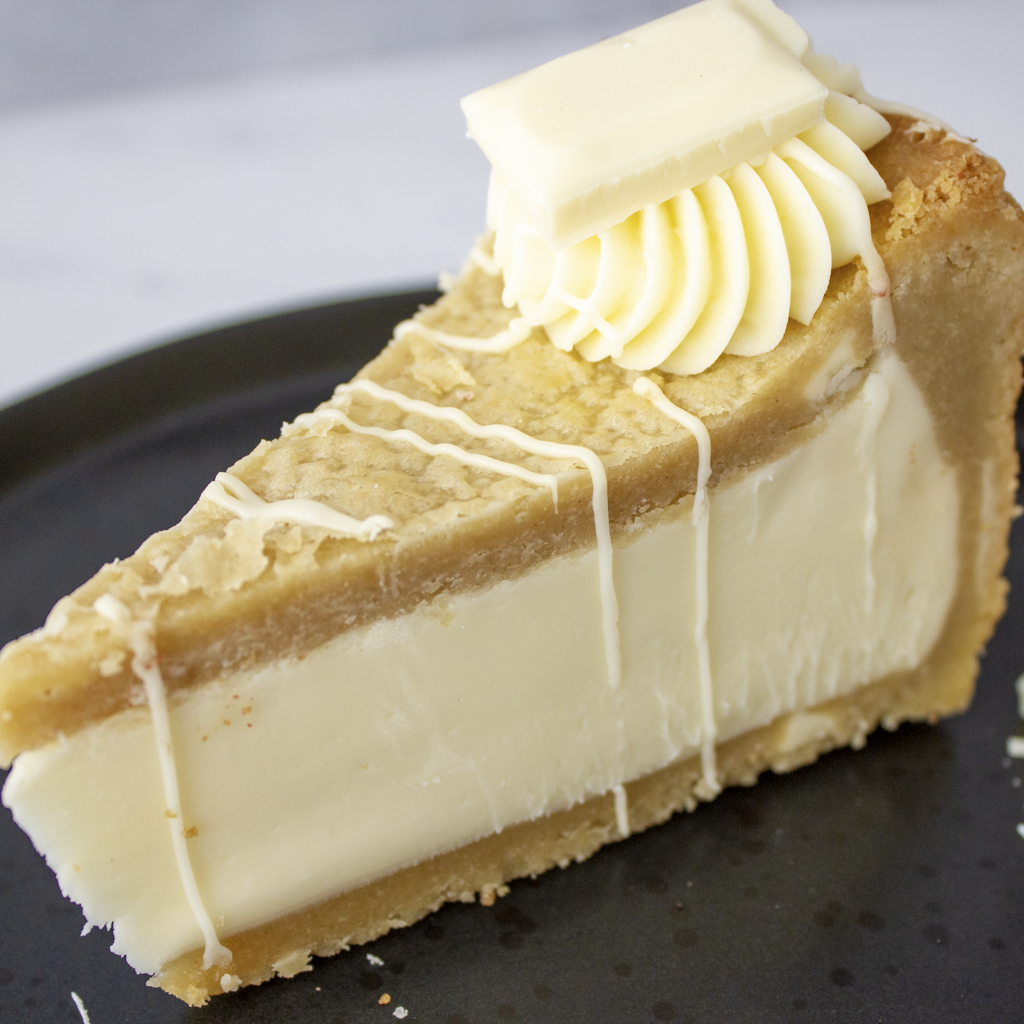 A close-up of a slice of white chocolate cookie pie on a dark plate, topped with a square of white chocolate and piped cream.