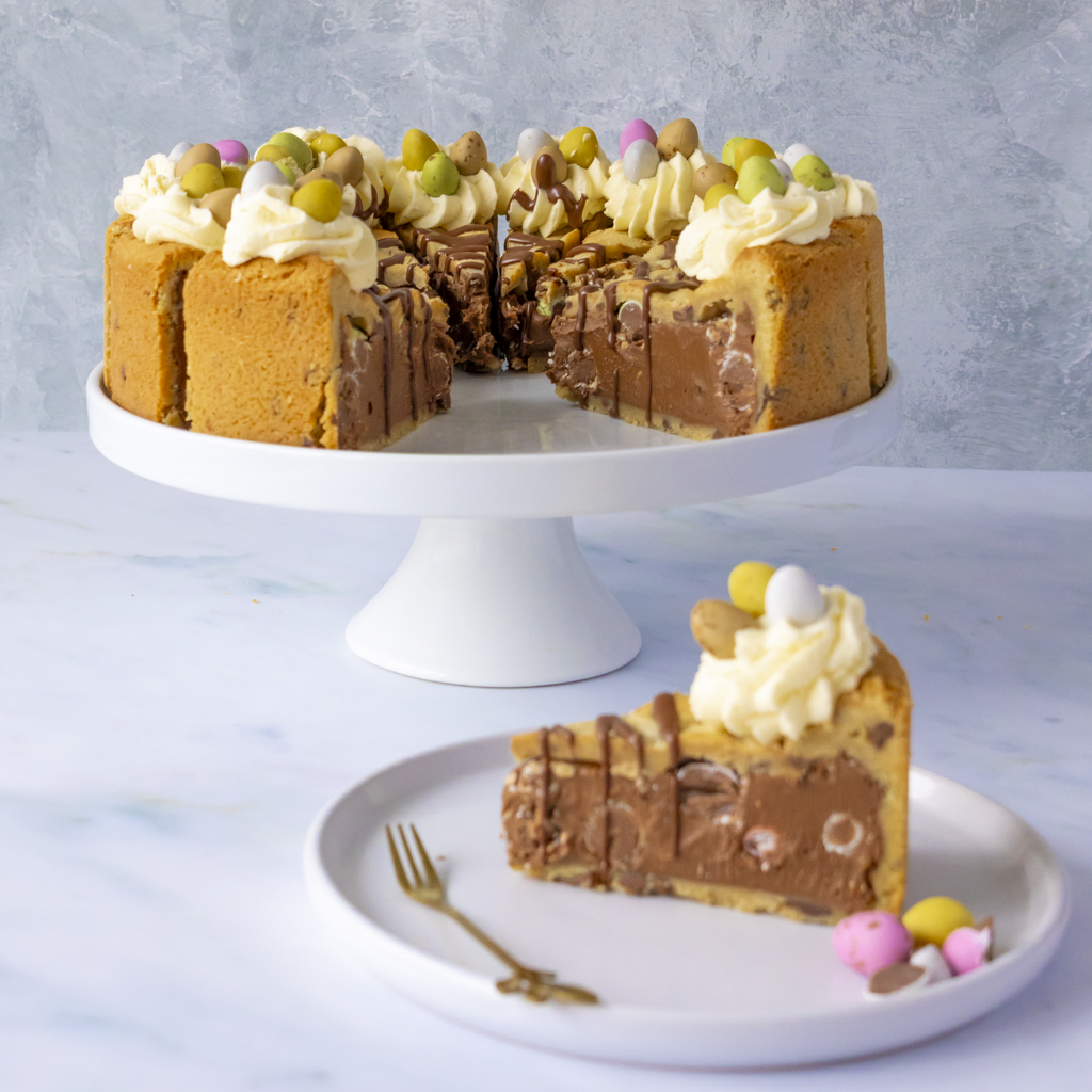 Multi-layered Easter Mini Egg cookie cake with a slice on a plate and a fork, against a light gray background.