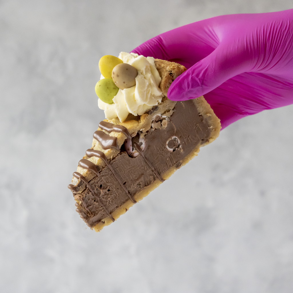 Slice of DoughGirl Mini Egg Cookie Pie Slice with Easter chocolate mini eggs held by a hand in a purple glove on a gray background.