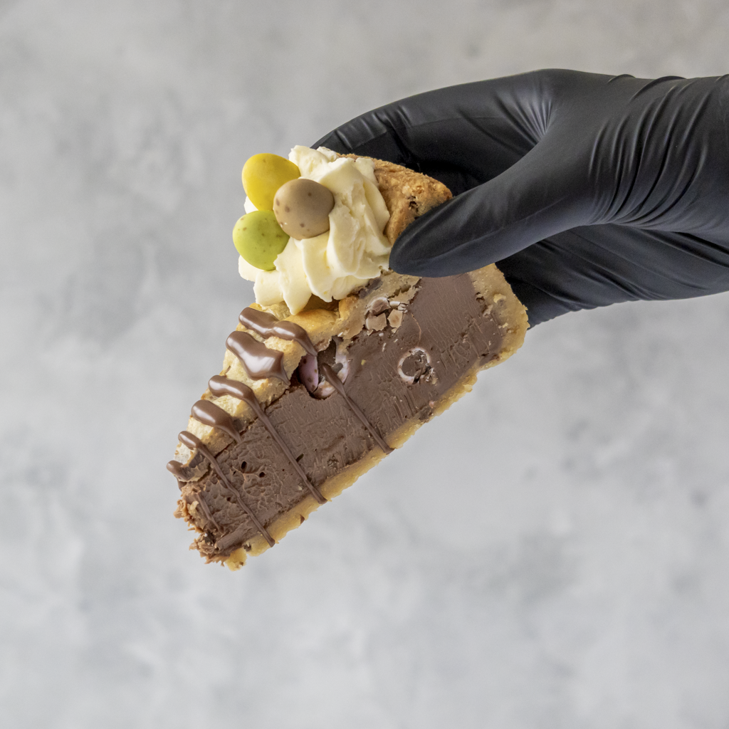 Slice of Limited Edition Mini Egg Cookie Pie with chocolate Mini Eggs held by a gloved hand on a gray background