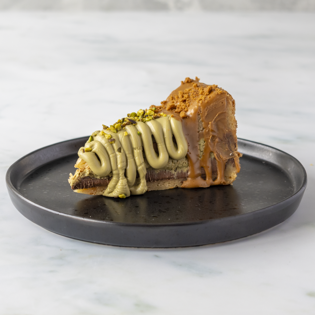 A slice of gourmet pastry on a dark round plate, featuring a chocolate base, caramel drizzle, pistachio cream, and chopped pistachios, set on a light marble surface.