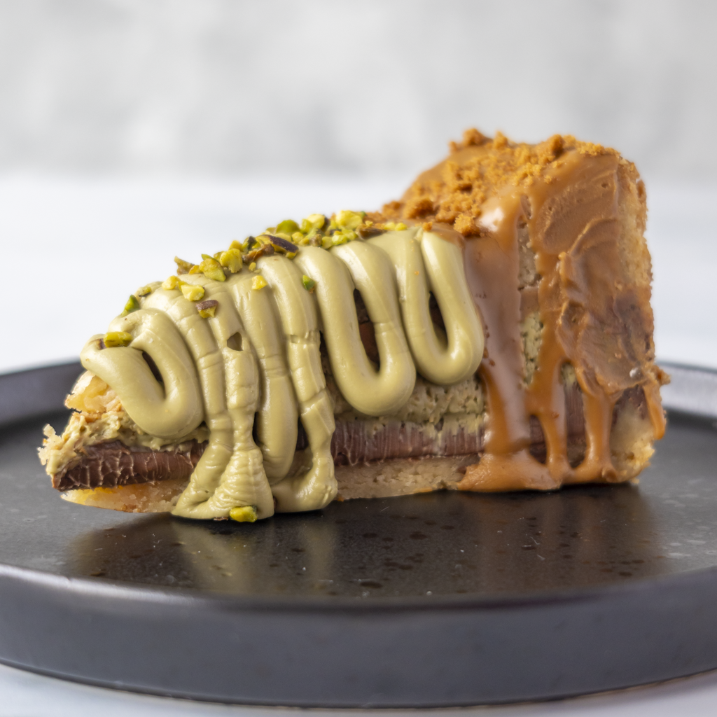 A close-up of a slice of Dubai Chocolate Cookie Pie with Biscoff on a dark round plate, featuring a biscuit base, chocolate layer, pistachio cream, caramel drizzle, and chopped pistachio topping.
