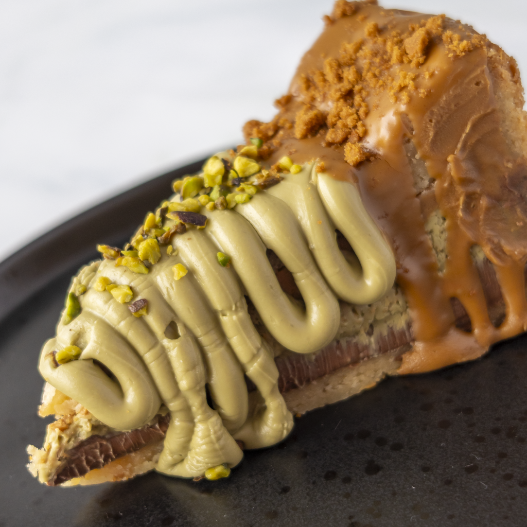 A close-up of a slice of dessert on a dark plate, featuring a light beige drizzle, a brown base, and chopped green pistachio pieces on top.