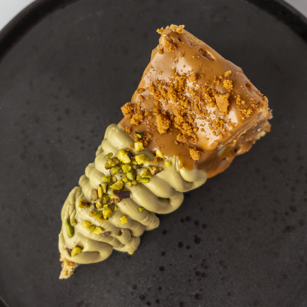 A close-up shot of a gourmet dessert slice, featuring a piece of chocolate cookie pie topped with caramel drizzle and cookie crumbs, dipped in pistachio-flavored cream and sprinkled with chopped pistachio nuts, served on a dark textured plate.