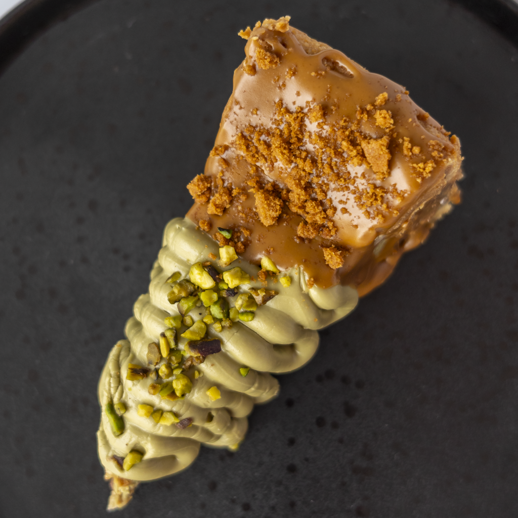 A close-up of a dessert slice on a dark plate, featuring a light green pistachio cream base topped with caramel brown glaze and crumbled cookie crumbs, with chopped green pistachios sprinkled on top.