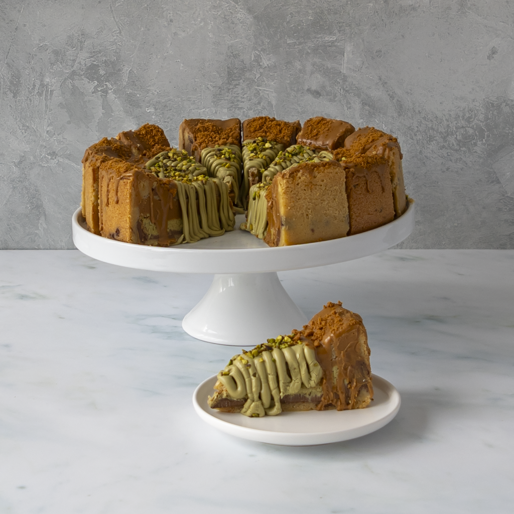 A sliced Dubai Chocolate Cookie Pie Slice with Biscoff with green pistachio drizzle and chopped pistachio topping, displayed on a white cake stand with a single slice on a small plate in front, against a gray textured wall and white marble surface.