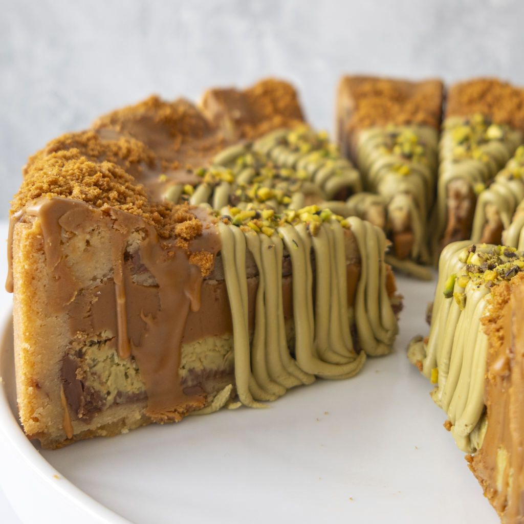 A close-up of a sliced layered dessert cookie pie on a white plate, featuring a biscuit or cookie base, chocolate and caramel drizzle, and pistachio filling with chopped pistachio on top.