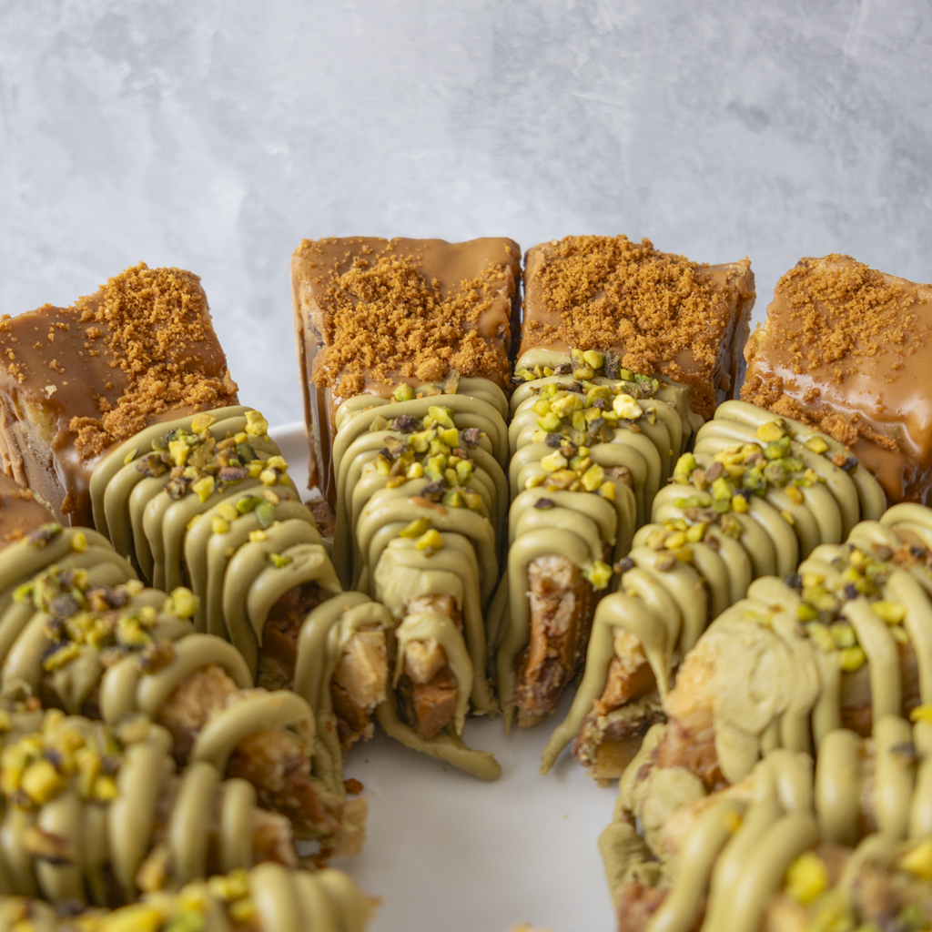 A close-up of several chocolate-dipped cookie pie slices arranged on a white plate against a light grey textured background. The pastries have a caramel-tan base topped with crumbled brown crumbs and drizzles of pale green chocolate, with chopped pistachio pieces on top.