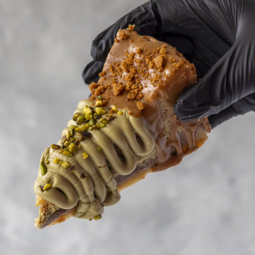 A close-up of a hand wearing a black glove holding a Dubai Chocolate Cookie Pie Slice with Biscoff with light brown drizzle, crumb topping, and pistachio pieces on a soft grey background.
