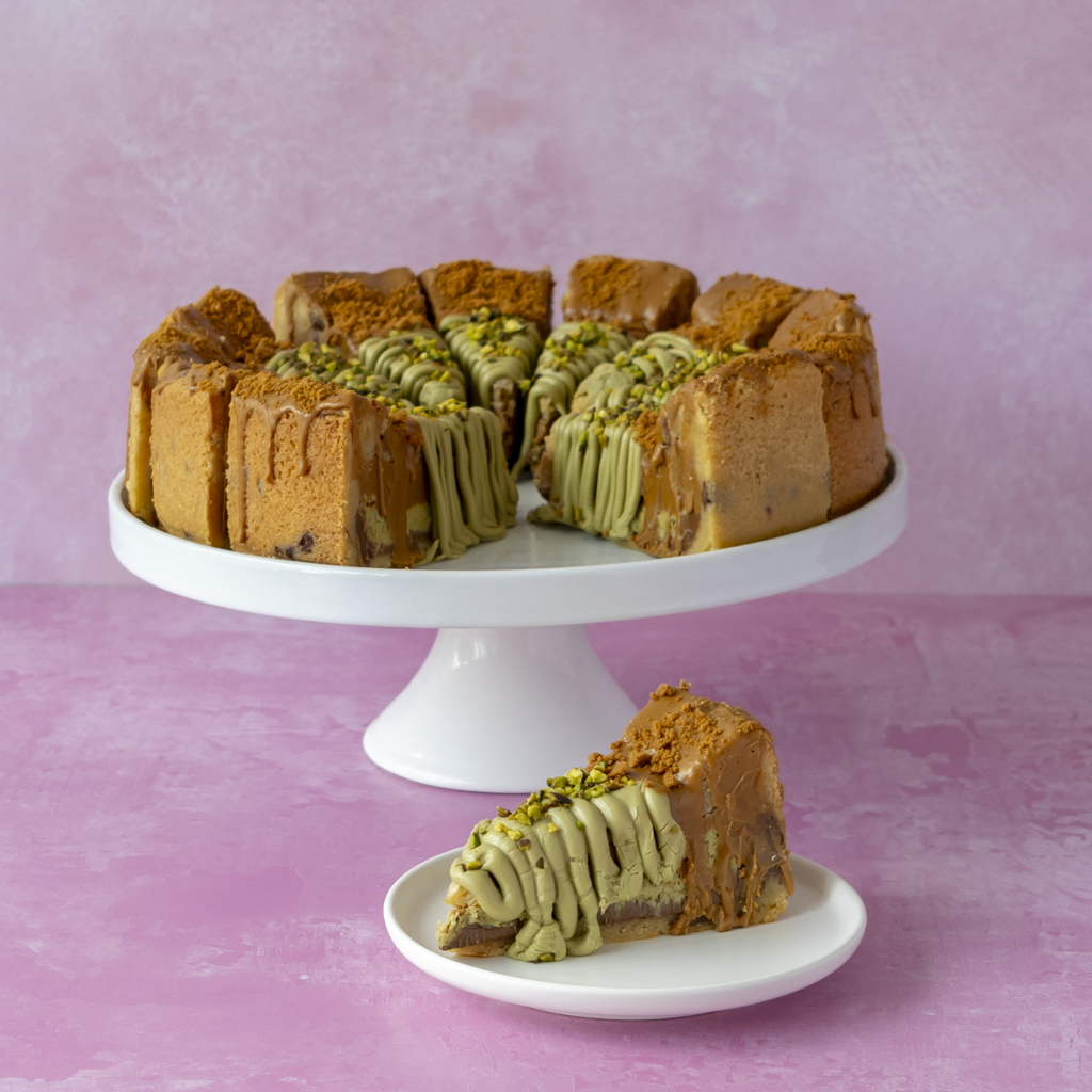 A round layered cookie pie on a white pedestal stand with a slice placed on a small white plate in front, set against a soft pink textured background.