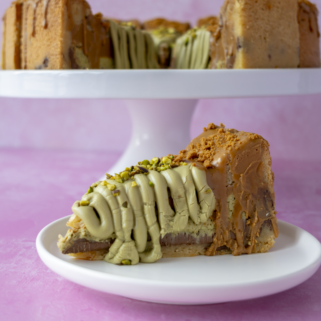 A slice of layered cookie pie on a white plate in the foreground, with a larger cake on a white pedestal stand in the background. The cookie pie slice has light green pistachio filling, a brown chocolate layer, and is topped with drizzle and chopped pistachio nuts.