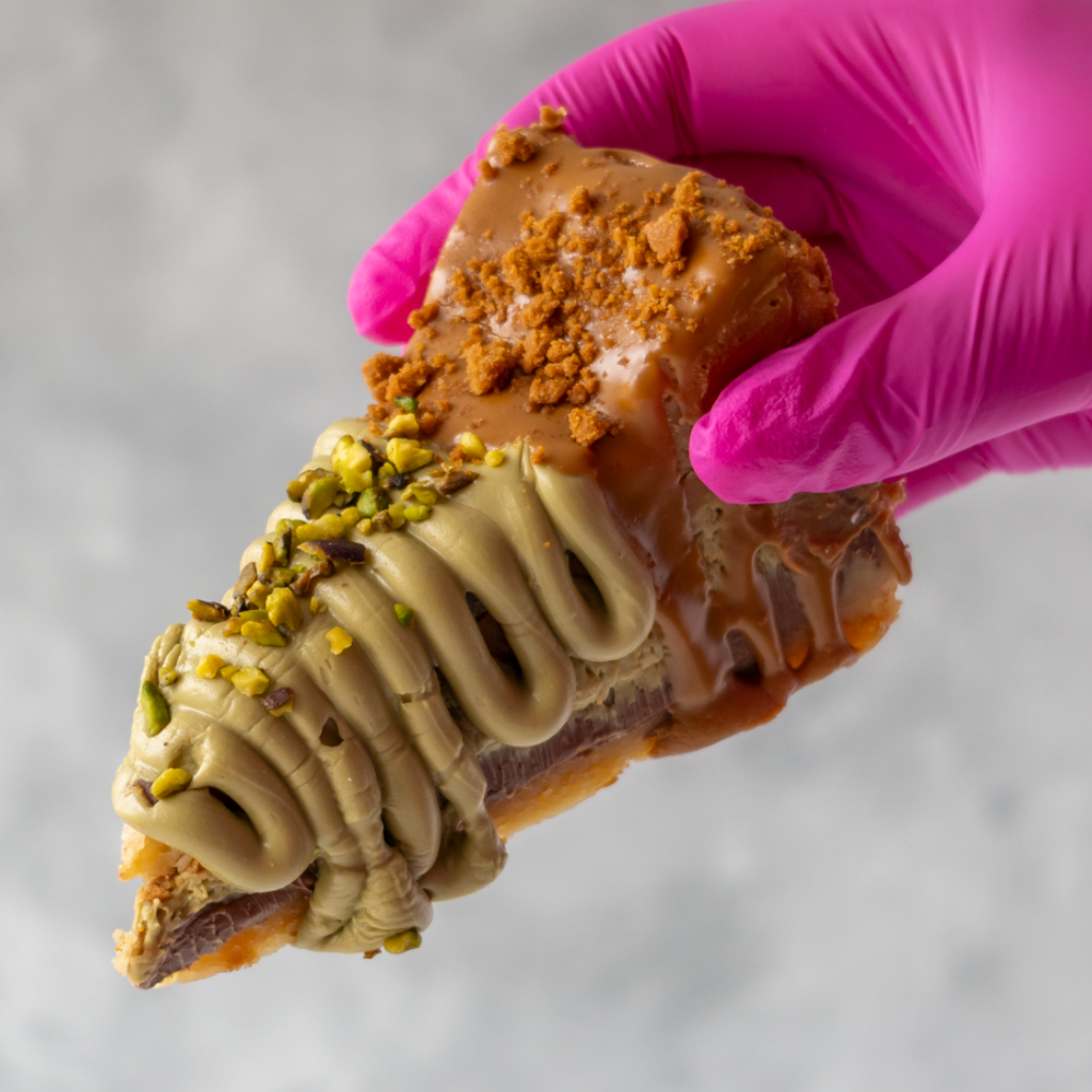 A close-up of a hand wearing a bright pink glove holding a Dubai Chocolate Cookie Pie Slice with Biscoff topped with light brown caramel, piped cream-colored frosting, chopped pistachio, and crumbled cookie pieces, against a soft light gray background.
