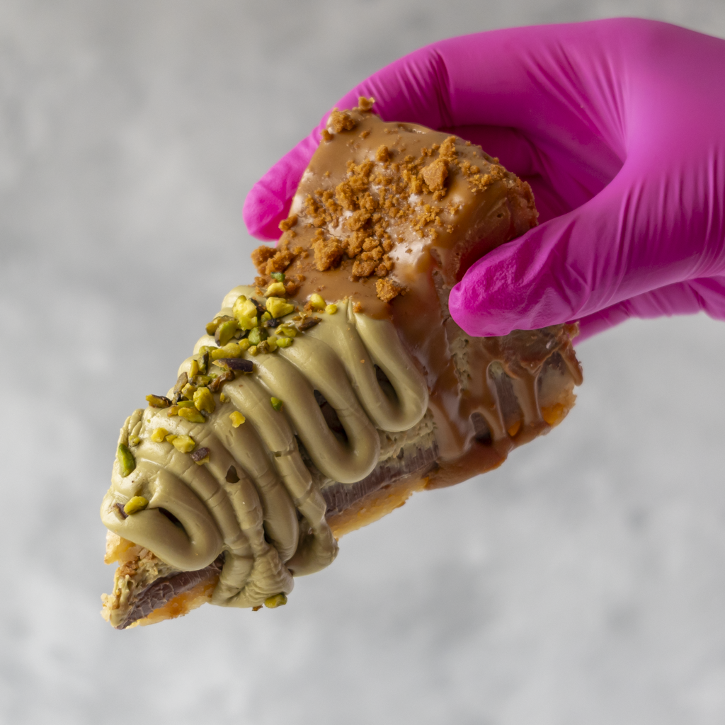 A close-up of a hand wearing a bright pink glove holding a pastry topped with light brown caramel, piped cream-colored frosting, chopped pistachio, and crumbled cookie pieces, against a soft light gray background.