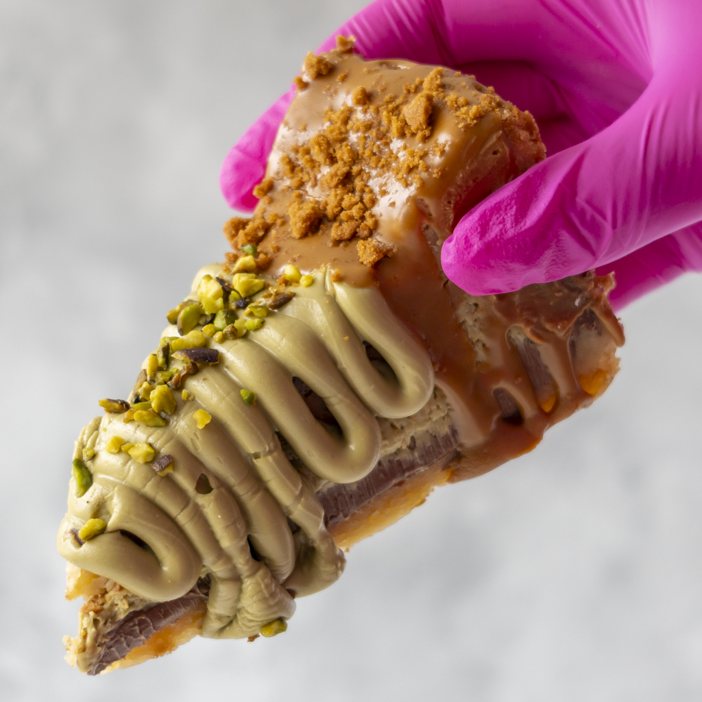 A close-up of a hand wearing a bright pink glove holding a pastry slice. The pastry has a glossy caramel-brown glaze, piped light beige cream, chopped pistachio pieces, and a crumb topping, set against a soft light gray background.