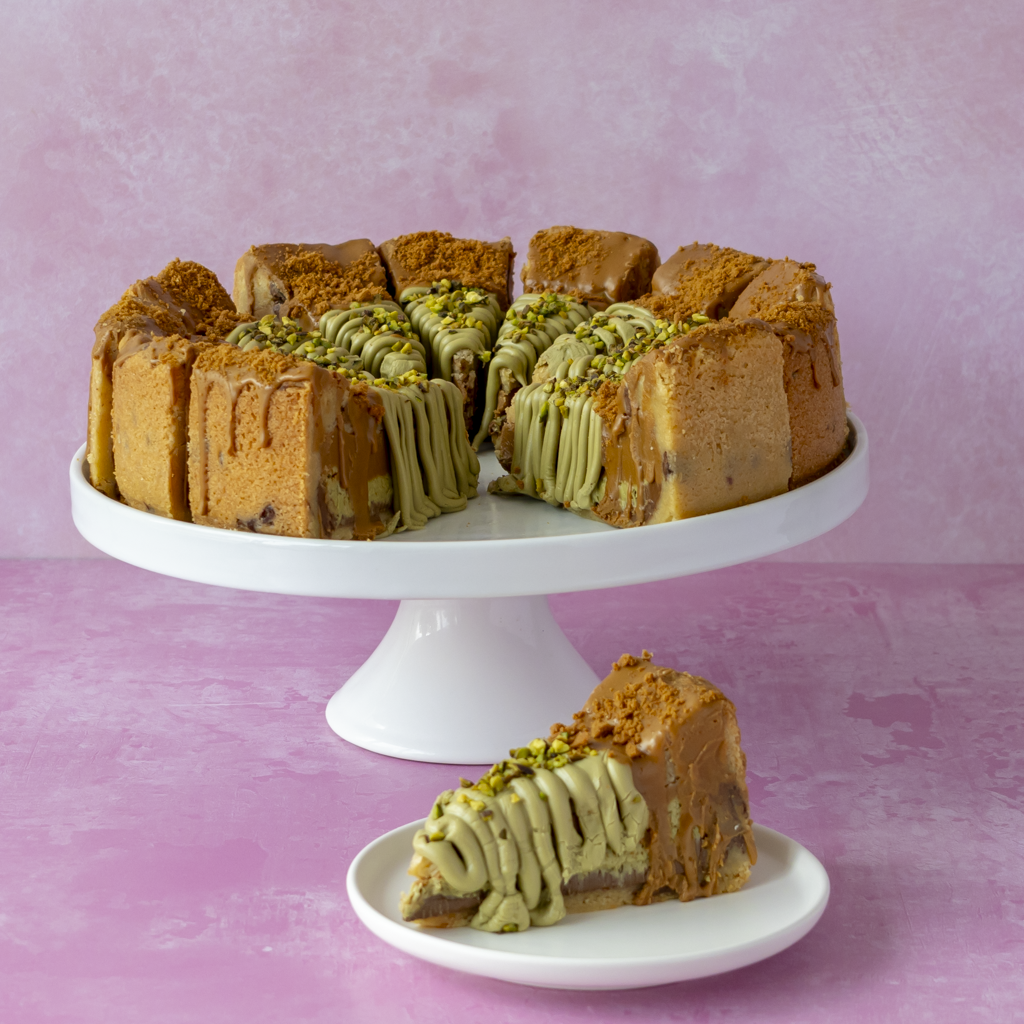 A slice of layered cookie pie with chocolate and pistachio layers, topped with green icing and chopped pistachio, displayed on a white pedestal cake stand with a single slice in front on a small plate against a pink background.