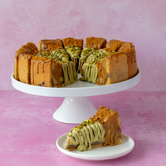 A slice of layered cookie pie with chocolate and pistachio layers, topped with green icing and chopped pistachio, displayed on a white pedestal cake stand with a single slice in front on a small plate against a pink background.