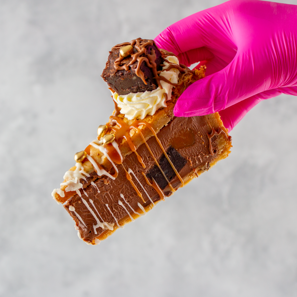 Hand holding a Salted Caramel Brownie Cookie Pie with chocolate, caramel, and whipped cream against a neutral background