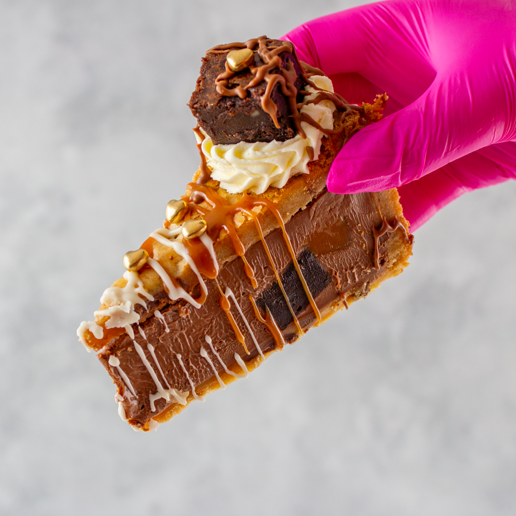 Hand holding a Limited Edition Salted Caramel Brownie Cookie Pie with golden hearts and chocolate drizzle against a neutral background