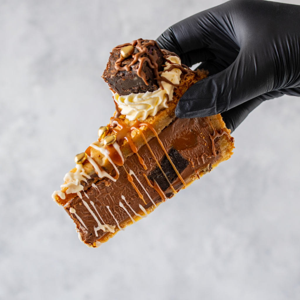 Hand holding a DoughGirl Salted Caramel Brownie Cookie Pie with chocolate, caramel, and white cream against a light background