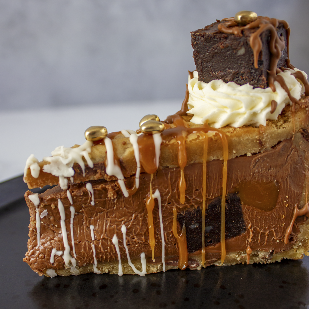 Close-up of a layered Salted Caramel Brownie Cookie Pie with chocolate, caramel, and golden heart on a dark surface.