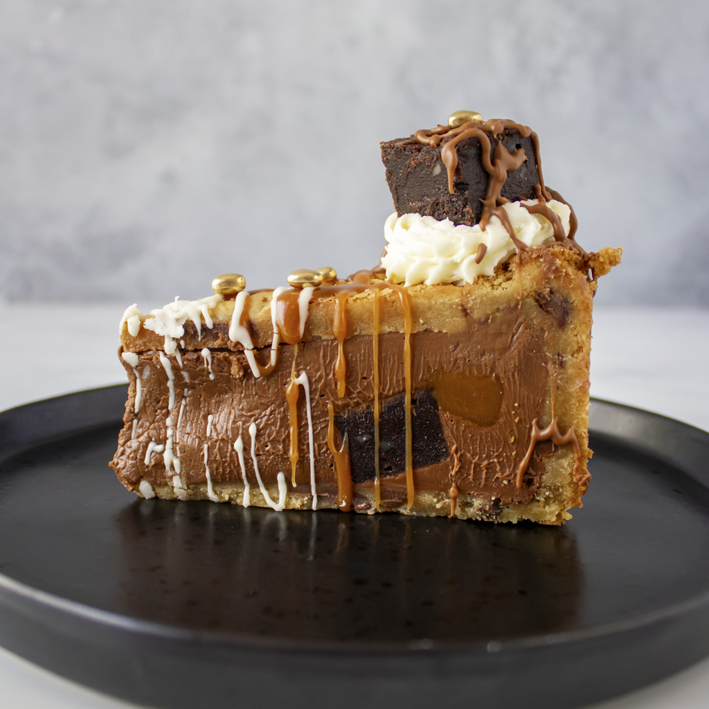 Slice of Salted Caramel Brownie Cookie Pie with chocolate and caramel on a black plate against a gray background