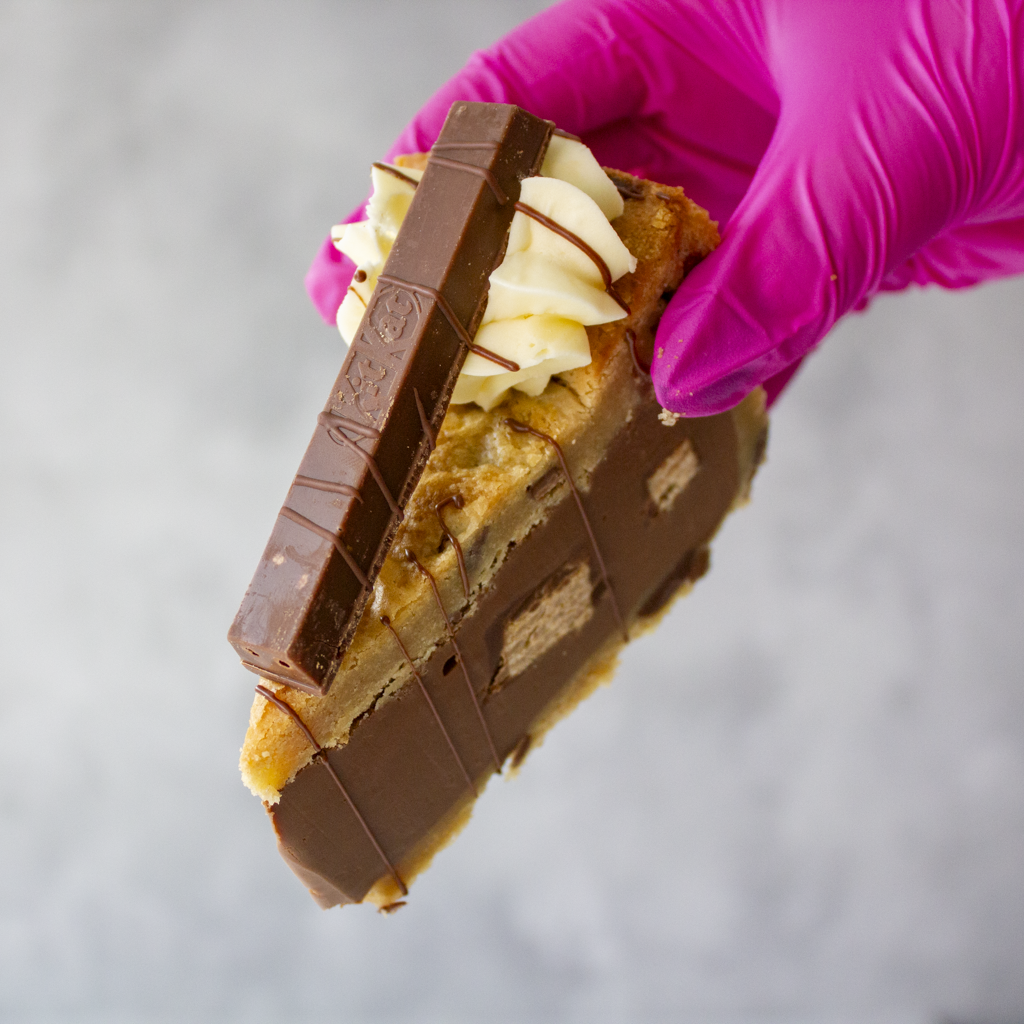 Hand holding a KitKat Crunch Cookie Pie by DoughGirl UK with a full KitKat chocolate bar, wearing a pink glove on a gray background
