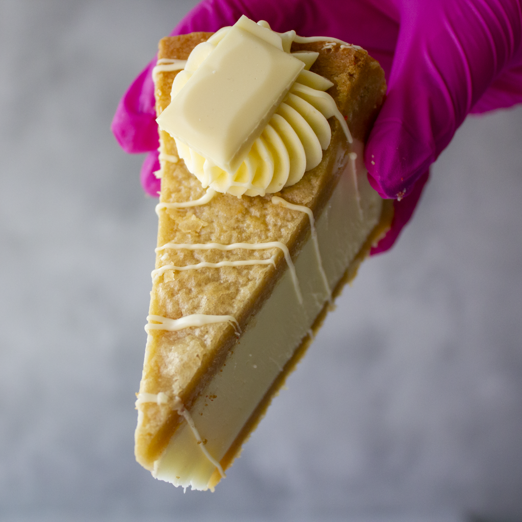 A close-up of a hand wearing a bright pink glove holding a slice of Cookie Pie with Milkybar with a dollop of whipped cream and white chocolate drizzle, set against a soft grey background.