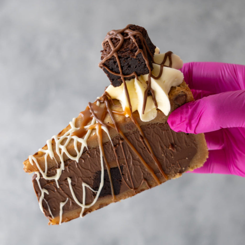 A close-up of a hand wearing a bright pink glove holding a slice of Caramel Brownie Cookie Pie Slice decorated with whipped cream, caramel drizzle, and a brownie on a grey background.