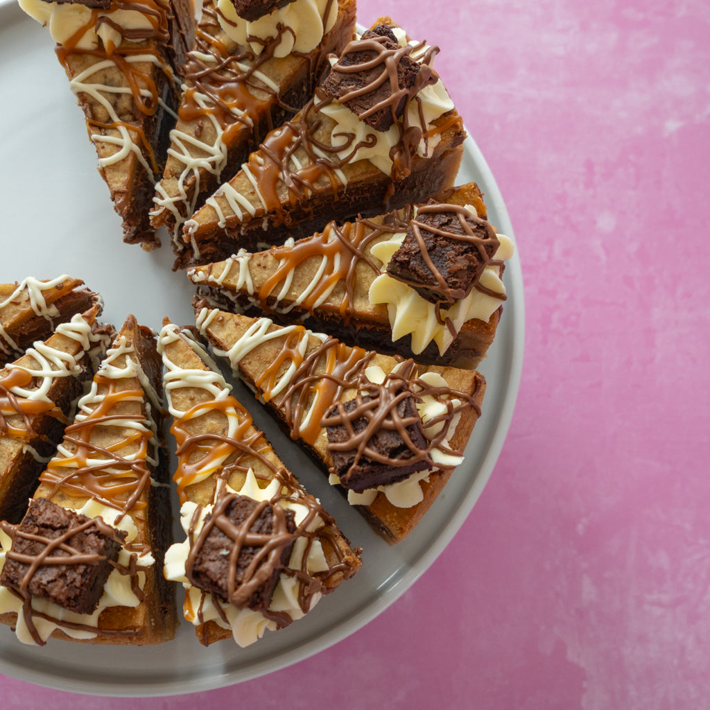 Slices of chocolate brownie cookie pie with caramel drizzle, white chocolate drizzle, and fresh brownie pieces, served on a light gray plate against a pink background.