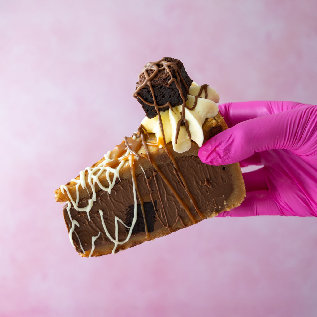 A slice of Caramel Brownie Cookie Pie Slice with whipped cream, drizzles, and a brownie topped with chocolate sauce, held by a hand wearing a bright pink glove against a soft pink background.