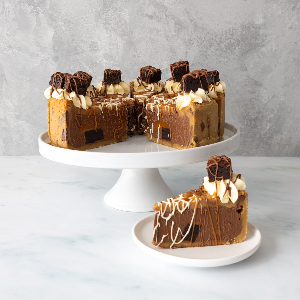 A full caramel brownie cookie pie with caramel drizzle and whipped cream sits on a white cake stand, with one slice placed on a small plate in front. The cookie pie has dark chocolate chunks and is topped with chocolate pieces, set against a light grey textured background.