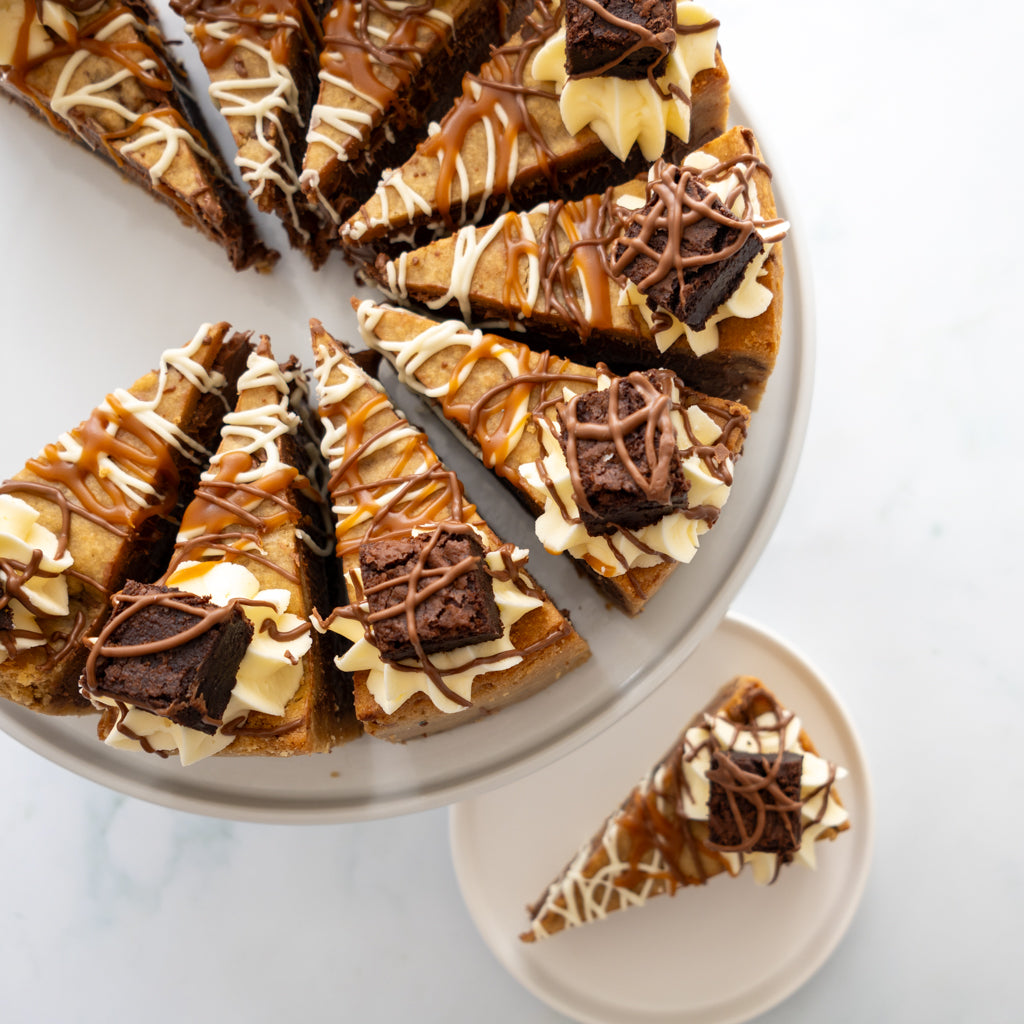 A sliced Full Caramel Brownie Cookie Pie with brownie, chocolate drizzle, caramel, and cream, presented on a white cake stand with one slice placed on a small plate below, on a light marble surface.